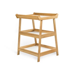 May include: A light-coloured wooden changing table with three levels. The top level has a raised edge, and the two lower levels have white surfaces. The table is made of light-coloured wood and has a simple, modern design.