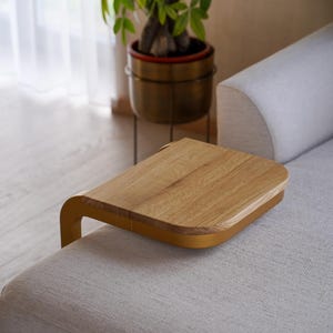 Op de afbeelding: Een houten sofa-armtafel met een licht eiken oppervlak en een gebogen, mosterdkleurig frame. De tafel is ontworpen om op de arm van een bank te rusten en een handig oppervlak te bieden. De achtergrond bevat een plant in een messing pot.