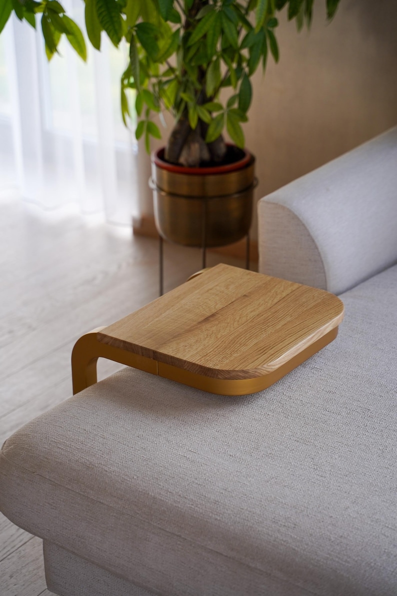 Op de afbeelding: Een houten sofa-armtafel met een licht eiken oppervlak en een gebogen, mosterdkleurig frame. De tafel is ontworpen om op de arm van een bank te rusten en een handig oppervlak te bieden. De achtergrond bevat een plant in een messing pot.