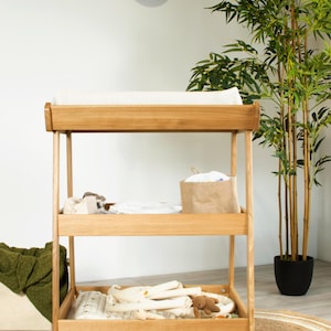 May include: Wooden baby changing unit with three shelves. The top shelf has a white changing mat. The middle shelf holds a brown paper bag and baby items. The bottom shelf has baby blankets and a teddy bear. A round jute rug is on the floor.