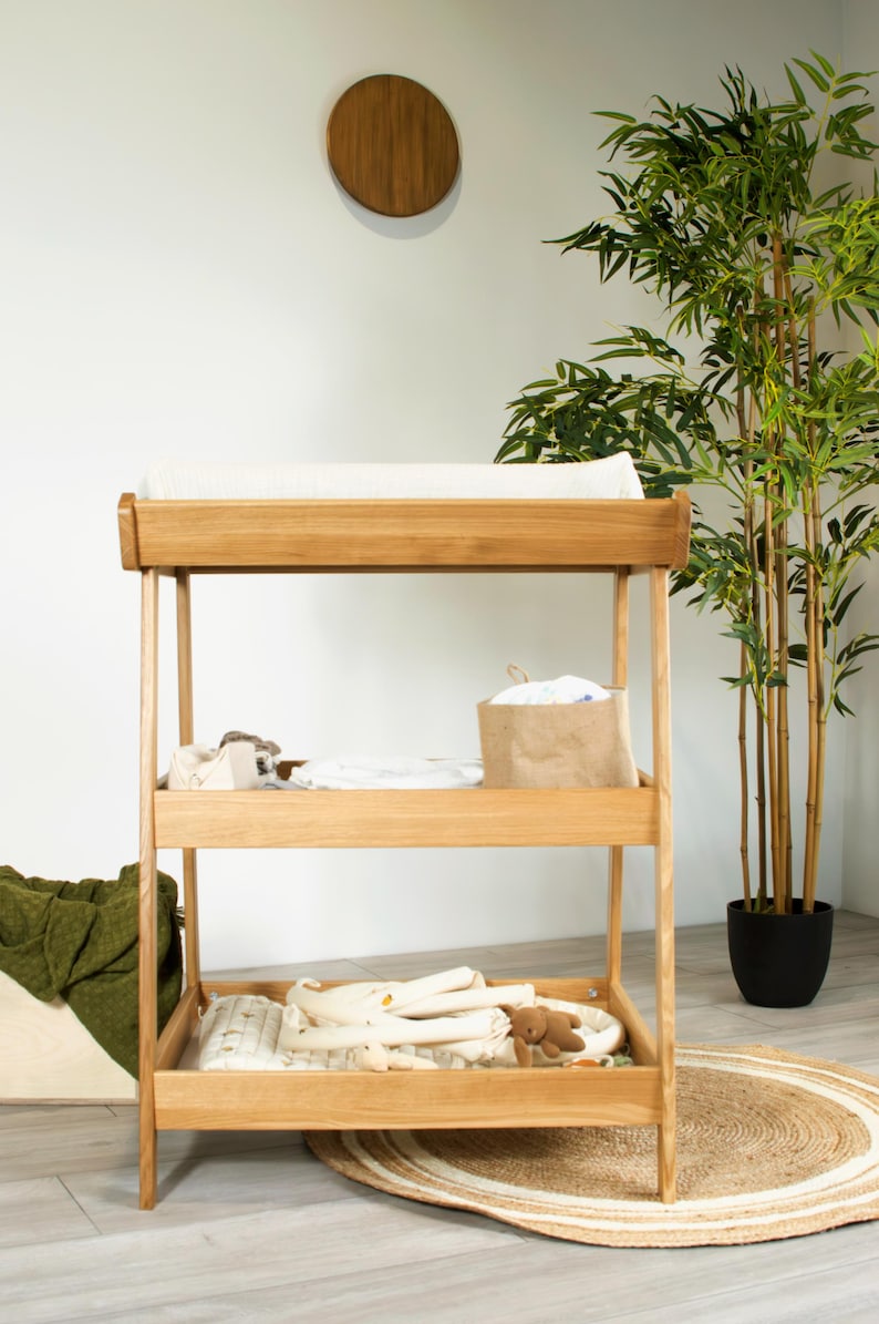 May include: Wooden baby changing unit with three shelves. The top shelf has a white changing mat. The middle shelf holds a brown paper bag and baby items. The bottom shelf has baby blankets and a teddy bear. A round jute rug is on the floor.