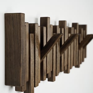 May include: A modern, wall-mounted coat rack made of dark brown wood. The design features vertical wooden slats of varying heights, with several angled hooks extending outward. The rack is mounted on a white wall.