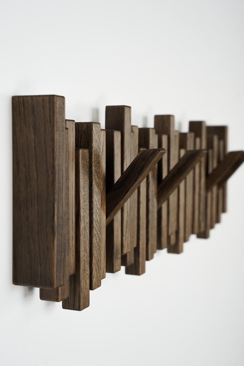 May include: A modern, wall-mounted coat rack made of dark brown wood. The design features vertical wooden slats of varying heights, with several angled hooks extending outward. The rack is mounted on a white wall.
