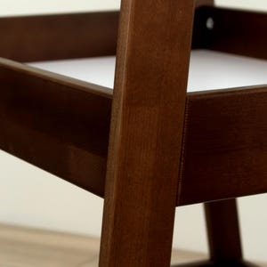 May include: Close-up of a wooden chair frame. The chair is constructed with dark brown wood, showcasing the grain. The design features a sturdy, angular structure with visible joints and screws. The background is a neutral off-white.