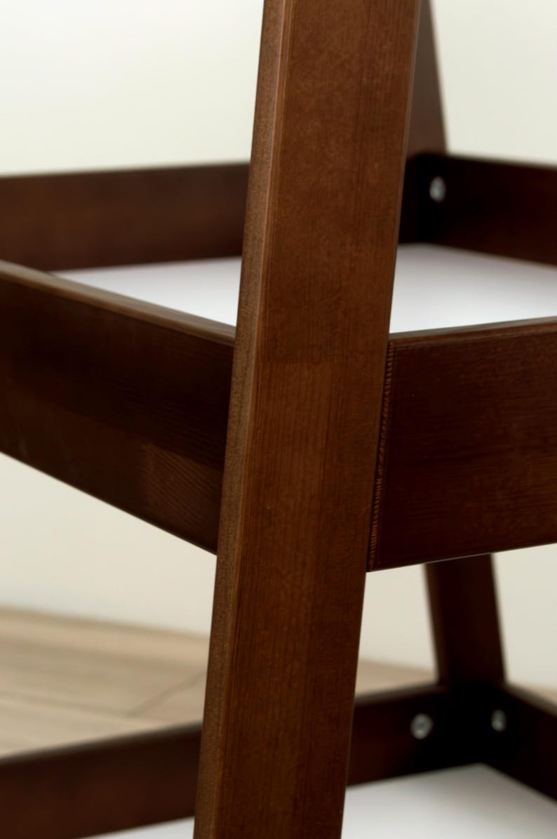 May include: Close-up of a wooden chair frame. The chair is constructed with dark brown wood, showcasing the grain. The design features a sturdy, angular structure with visible joints and screws. The background is a neutral off-white.