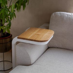 Op de afbeelding: Een houten bijzettafel met een lichte eiken afwerking en een wit metalen frame is bevestigd aan de arm van een beige bank. Een potplant met groene bladeren staat op de achtergrond.