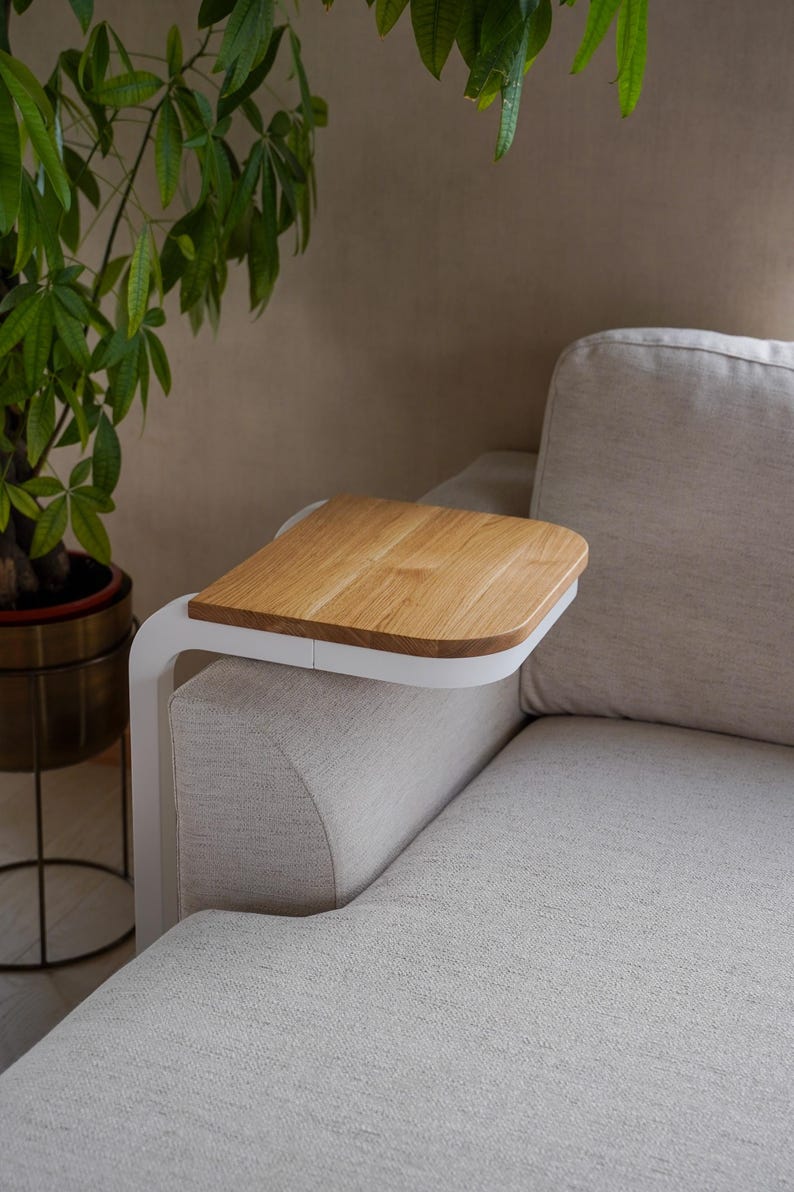 Op de afbeelding: Een houten bijzettafel met een lichte eiken afwerking en een wit metalen frame is bevestigd aan de arm van een beige bank. Een potplant met groene bladeren staat op de achtergrond.