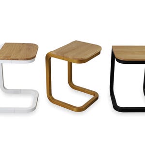Op de afbeelding: Drie moderne bijzettafels met een C-vormige basis en een rechthoekig houten blad. E&eacute;n tafel heeft een witte basis, &eacute;&eacute;n is goudkleurig en de derde is zwart. De houten bladen hebben een natuurlijke houtnerf.
