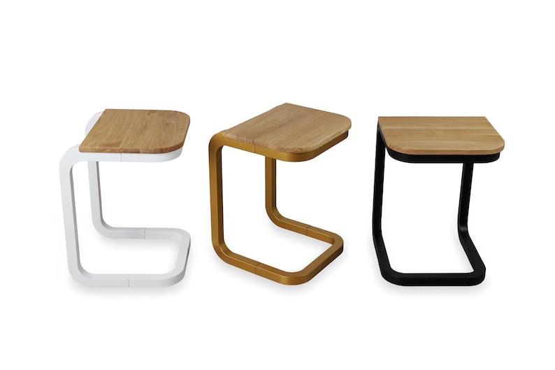 Op de afbeelding: Drie moderne bijzettafels met een C-vormige basis en een rechthoekig houten blad. E&eacute;n tafel heeft een witte basis, &eacute;&eacute;n is goudkleurig en de derde is zwart. De houten bladen hebben een natuurlijke houtnerf.