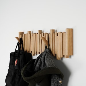May include: A wooden wall-mounted coat rack with a unique design. The rack features vertical wooden slats of varying heights, with two wooden hooks. A black backpack and a dark green coat with a hood are hanging on the hooks.