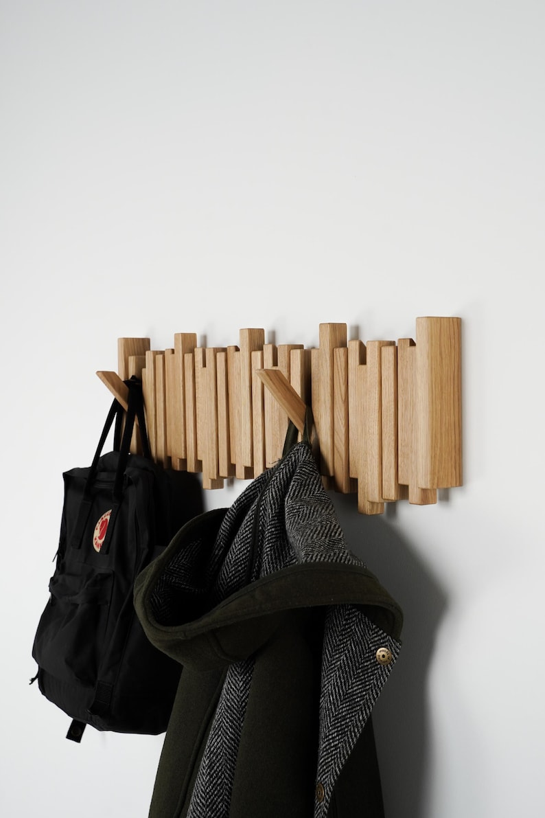 May include: A wooden wall-mounted coat rack with a unique design. The rack features vertical wooden slats of varying heights, with two wooden hooks. A black backpack and a dark green coat with a hood are hanging on the hooks.