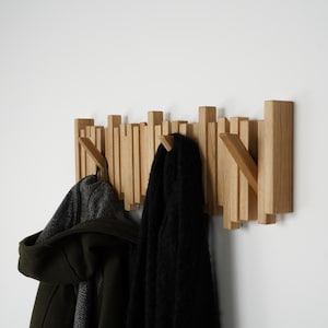 May include: Wooden wall-mounted coat rack with a unique design. The rack features multiple vertical wooden slats of varying heights, creating an interesting visual texture. A dark green coat and a black scarf hang from the hooks.