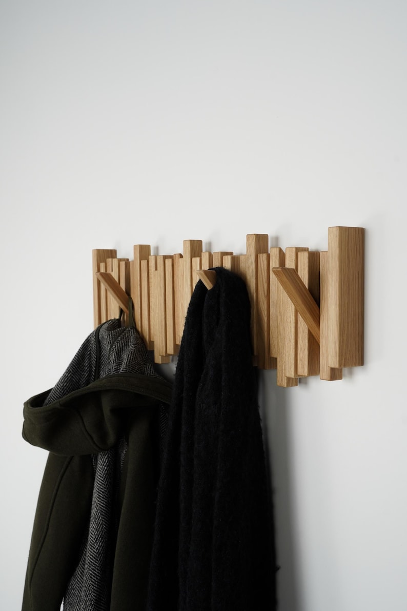 May include: Wooden wall-mounted coat rack with a unique design. The rack features multiple vertical wooden slats of varying heights, creating an interesting visual texture. A dark green coat and a black scarf hang from the hooks.