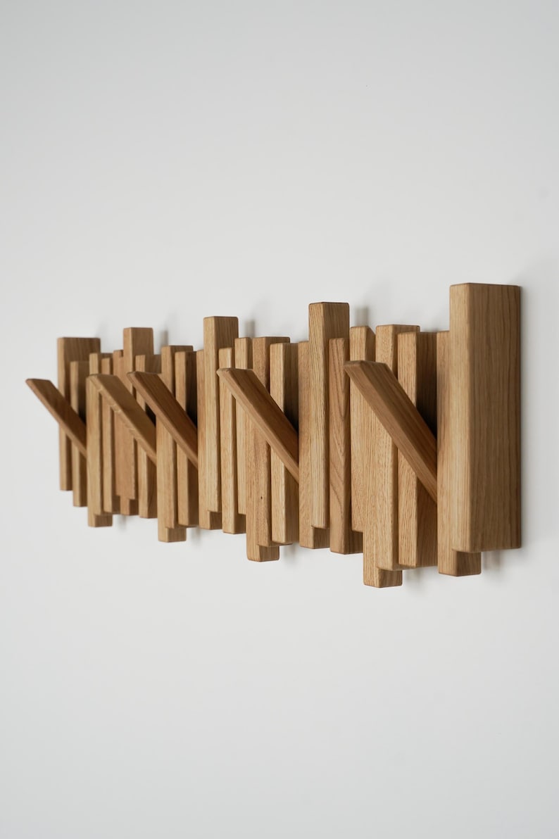 May include: Wooden wall-mounted coat rack with a modern design. The rack features multiple vertical wooden slats of varying heights, with angled hooks for hanging items. The natural wood has a light brown color, and the rack is mounted on a white wall.