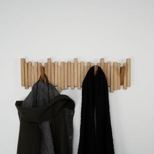 May include: A wooden coat rack with multiple vertical wooden pegs. A dark green coat with a hood and a black scarf hang from the rack. The rack is mounted on a white wall.
