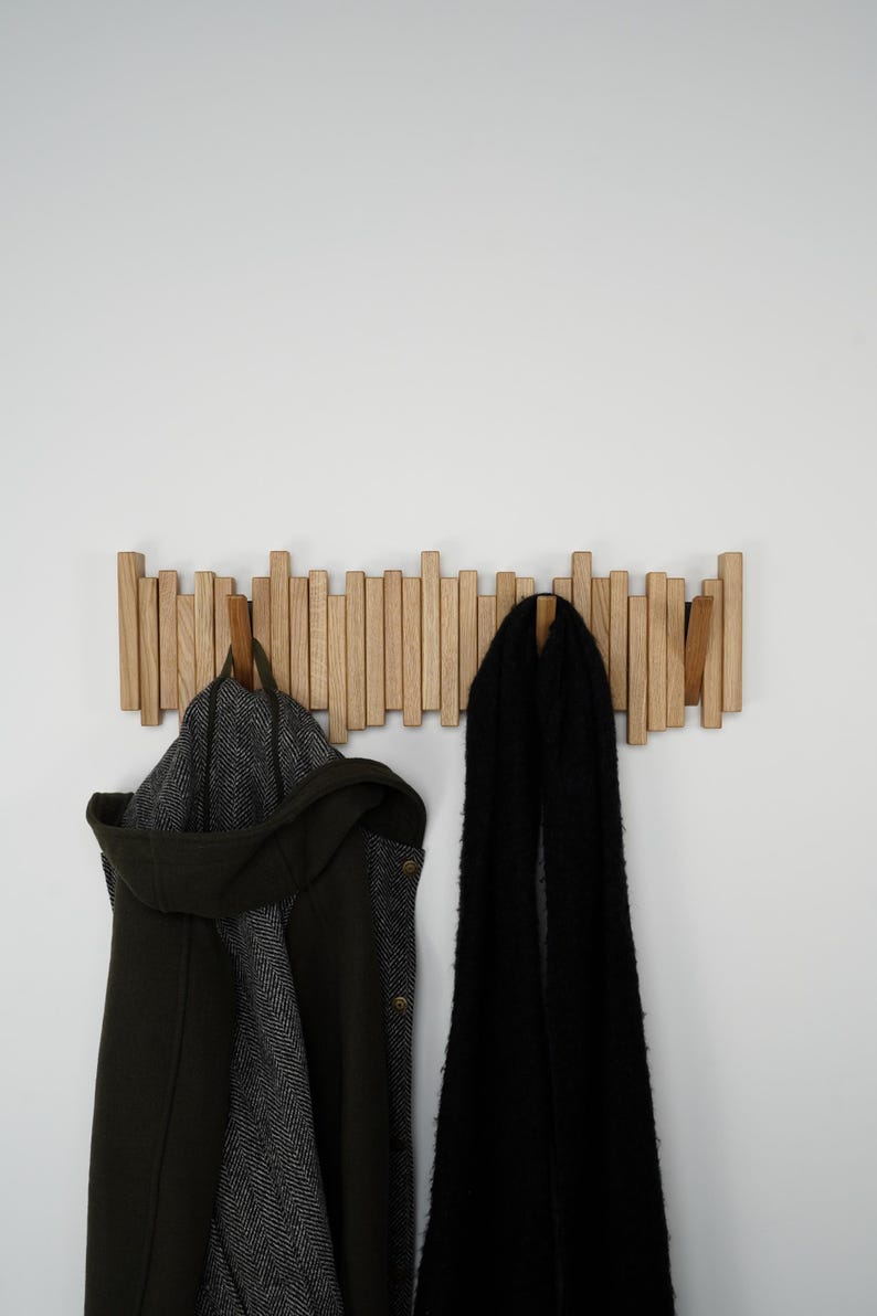 May include: A wooden coat rack with multiple vertical wooden pegs. A dark green coat with a hood and a black scarf hang from the rack. The rack is mounted on a white wall.