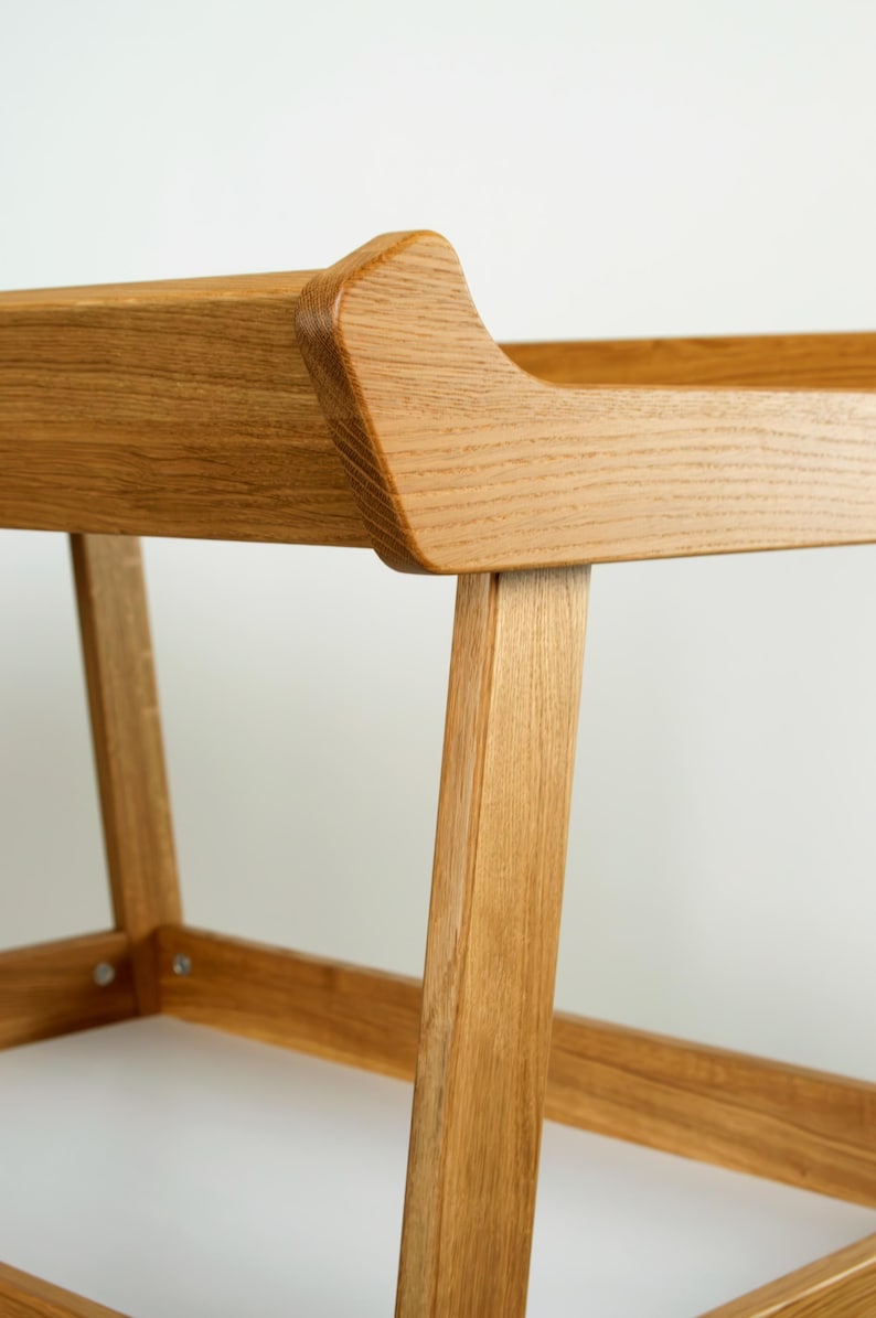 May include: Close-up of a wooden chair with a light brown finish. The chair has a curved backrest and angled legs, showcasing a minimalist design. The wood grain is visible, adding texture to the piece. The chair appears sturdy and well-crafted.