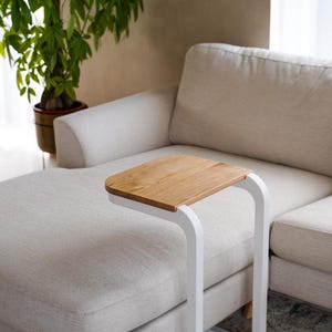 Op de afbeelding: Een moderne bijzettafel met een licht houten blad en een wit metalen frame. De tafel is ontworpen om over de arm van een bank te schuiven. De afmetingen van de tafel zijn ongeveer 51 cm lang, 30 cm breed en 61 cm hoog.