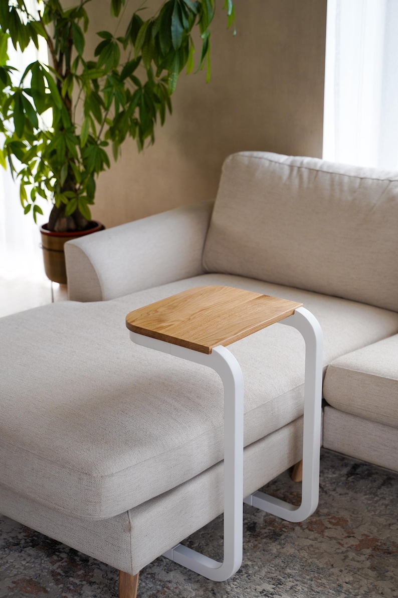 Op de afbeelding: Een moderne bijzettafel met een licht houten blad en een wit metalen frame. De tafel is ontworpen om over de arm van een bank te schuiven. De afmetingen van de tafel zijn ongeveer 51 cm lang, 30 cm breed en 61 cm hoog.