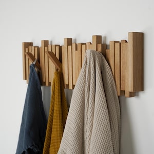 May include: A wooden wall-mounted coat rack with a series of vertical wooden pegs. Three items hang from the pegs: a navy blue towel, a mustard yellow towel, and a beige waffle-weave robe. The rack is mounted on a white wall.