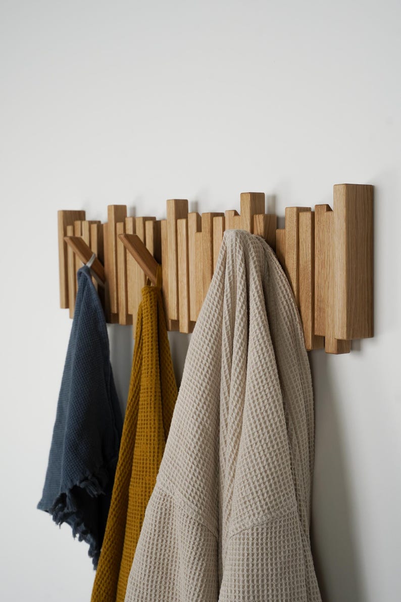 May include: A wooden wall-mounted coat rack with a series of vertical wooden pegs. Three items hang from the pegs: a navy blue towel, a mustard yellow towel, and a beige waffle-weave robe. The rack is mounted on a white wall.
