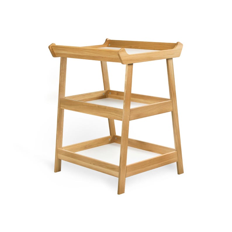 May include: A light-colored wooden changing table with three levels. The top level has a raised edge, and the two lower levels are open shelves. The table is made of light-colored wood and has a simple, modern design.