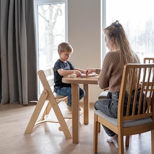 May include: A light wood children's table and chair set. The table is round, and the chairs are made of wood with a light gray cushion. A woman and a child are seated at the table.