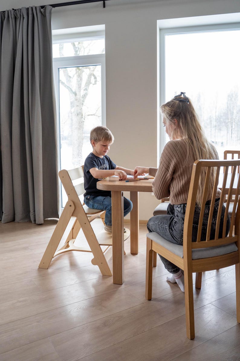 May include: A light wood children's table and chair set. The table is round, and the chairs are made of wood with a light gray cushion. A woman and a child are seated at the table.