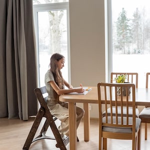 May include: A light wood dining table with a person sitting at it, writing in a notebook. The table is surrounded by wooden chairs with gray cushions. A folding wooden chair is in the foreground. A plant sits on the table.