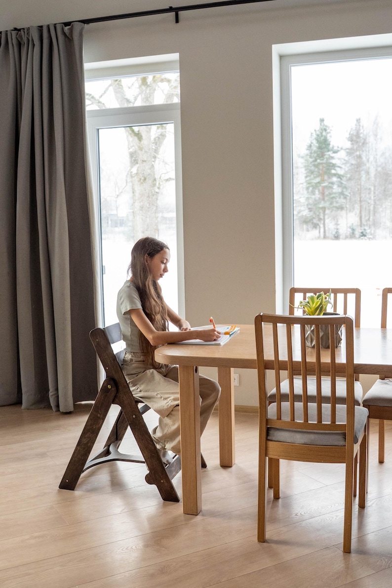May include: A light wood dining table with a person sitting at it, writing in a notebook. The table is surrounded by wooden chairs with gray cushions. A folding wooden chair is in the foreground. A plant sits on the table.