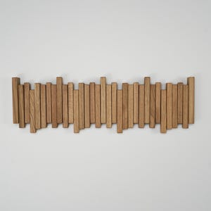 May include: A wooden wall-mounted coat rack with a series of vertical wooden slats of varying heights. The natural wood has a light brown color and is mounted on a white wall. The rack is designed for hanging coats or other items.
