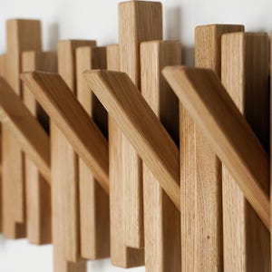 May include: Wooden wall-mounted coat rack with a modern design. The rack features multiple rectangular pegs of varying heights and angled hooks, all made from light-colored wood. The pegs are arranged in a staggered pattern, creating a visually interesting and functional piece.