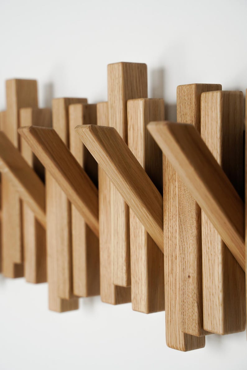 May include: Wooden wall-mounted coat rack with a modern design. The rack features multiple rectangular pegs of varying heights and angled hooks, all made from light-colored wood. The pegs are arranged in a staggered pattern, creating a visually interesting and functional piece.