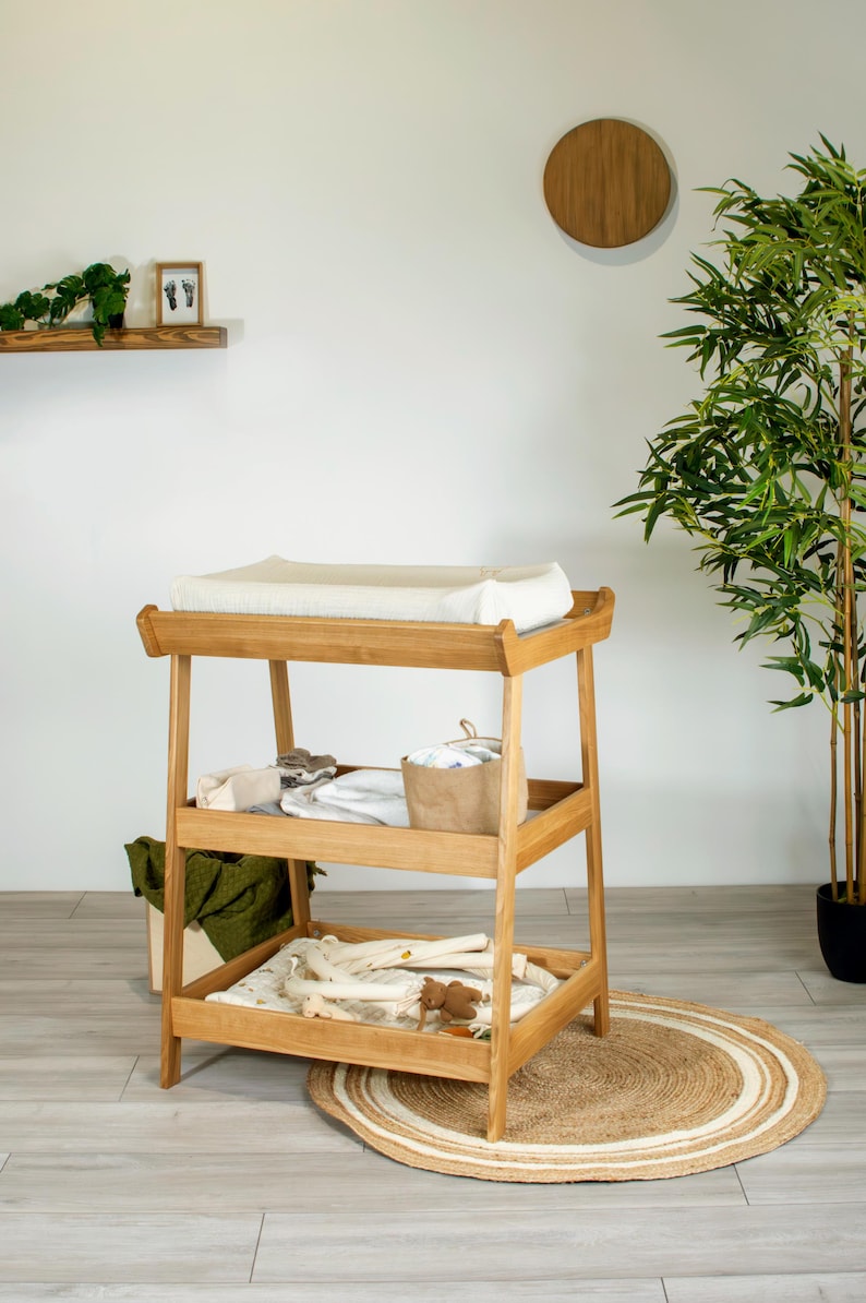 May include: A light wood baby changing table with three shelves. The top shelf has a white changing pad. The middle shelf holds a basket and folded items. The bottom shelf has baby clothes and toys. A round rug is beneath the table.