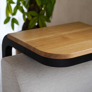 Op de afbeelding: Een moderne bijzettafel met een licht houten blad en een zwart gebogen frame. De tafel staat op een lichtgekleurde, gestoffeerde basis, met een groene plant op de achtergrond. De afmetingen van de tafel zijn niet zichtbaar.