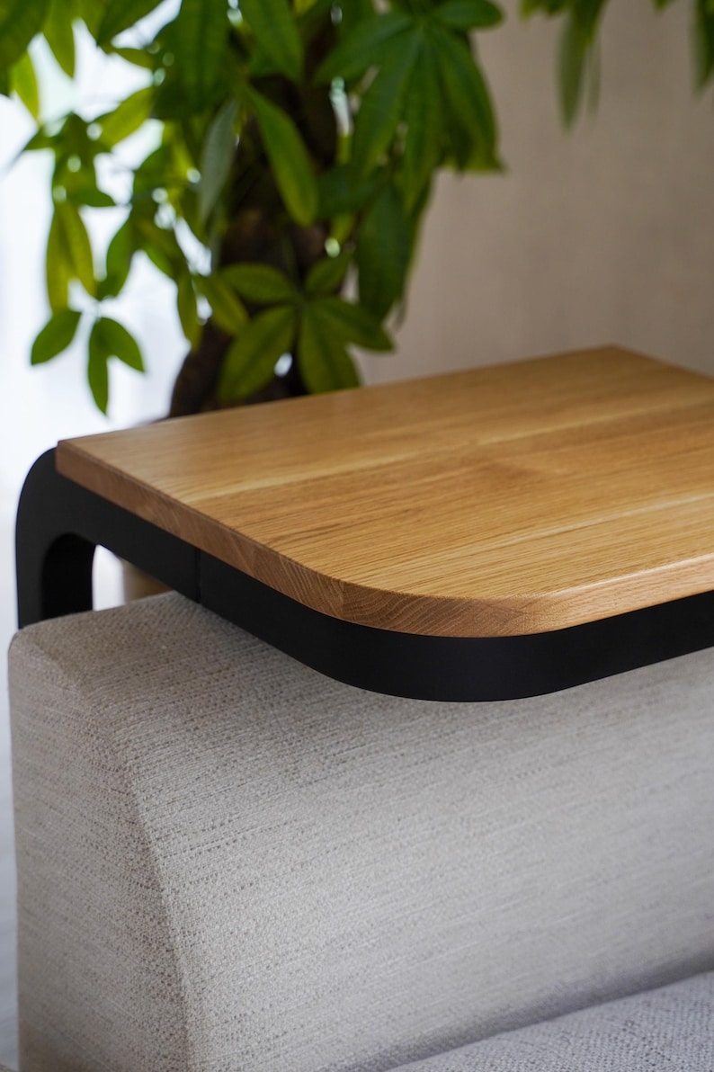 Op de afbeelding: Een moderne bijzettafel met een licht houten blad en een zwart gebogen frame. De tafel staat op een lichtgekleurde, gestoffeerde basis, met een groene plant op de achtergrond. De afmetingen van de tafel zijn niet zichtbaar.