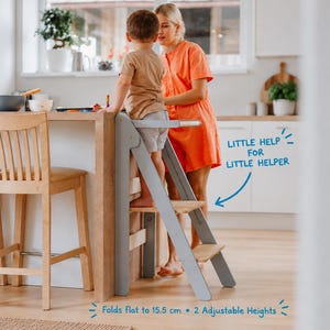 Könnte beinhalten: Ein hellgrauer Küchenhelfer-Tritt aus Holz mit zwei Stufen und einem Sicherheitsbügel. Der Text an der Seite lautet: Little help for little helper. Unten steht: Folds flat to 15.5 cm • 2 Adjustable Heights. Der Tritt steht an einer Küchentheke.