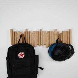 May include: A wooden wall-mounted coat rack with multiple vertical wooden dowels. A black backpack with a red and white logo and a black and blue bicycle helmet are hanging from the rack.