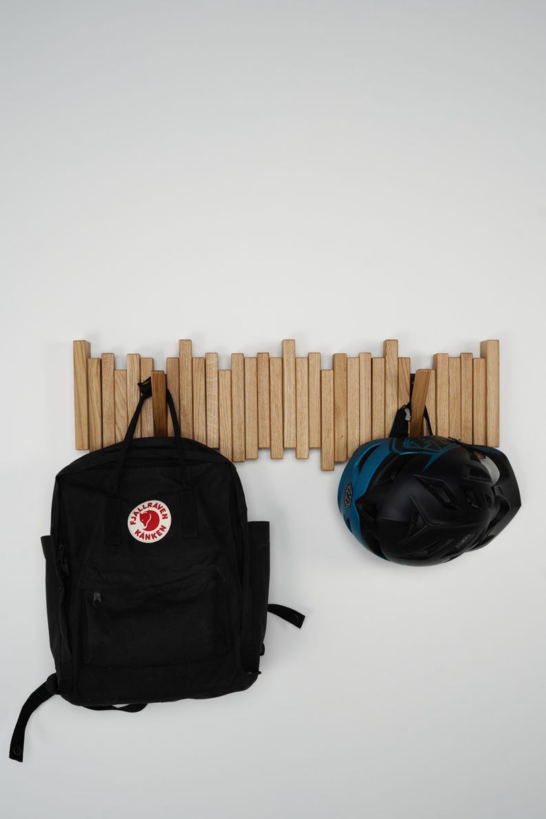 May include: A wooden wall-mounted coat rack with multiple vertical wooden dowels. A black backpack with a red and white logo and a black and blue bicycle helmet are hanging from the rack.