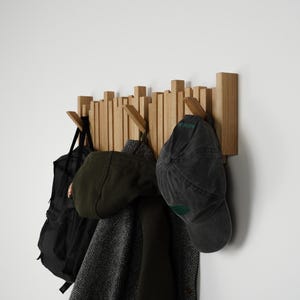 May include: Wooden wall-mounted coat rack with multiple pegs. A black backpack, a green cap, a dark gray cap, and a dark coat hang from the pegs. The rack is made of light-colored wood and is mounted on a white wall.