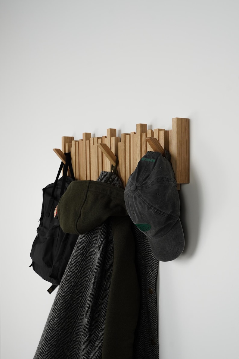May include: Wooden wall-mounted coat rack with multiple pegs. A black backpack, a green cap, a dark gray cap, and a dark coat hang from the pegs. The rack is made of light-colored wood and is mounted on a white wall.