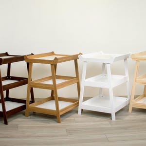 May include: Four wooden side tables in different colours: dark brown, natural wood, white, and light wood. Each table has a tray-like top and two shelves, with a simple, modern design. The tables are suitable for various home decor styles.