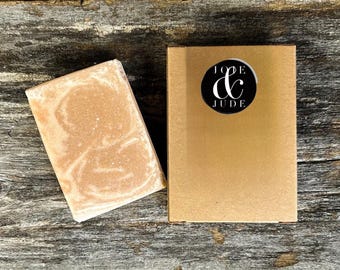 Puede incluir: Una pastilla de jabón marrón claro con un diseño veteado junto a una caja dorada. La caja tiene una pegatina circular en blanco y negro con las palabras "JOE & JUDE". El jabón y la caja están sobre una superficie de madera envejecida.