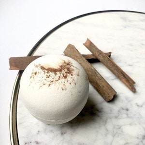 May include: A white bath bomb with a brown speckled design, alongside three cinnamon sticks. The bath bomb is on a marble surface, with a gold-coloured rim in the background. The image is well-lit and focuses on the bath bomb and cinnamon.
