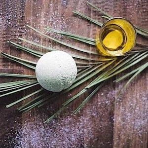 Pode incluir: Uma bomba de banho branca e um pequeno copo de líquido amarelo repousam sobre uma cama de folhas verdes, com cristais de sal espalhados sobre uma superfície de madeira. A bomba de banho tem uma superfície texturizada.