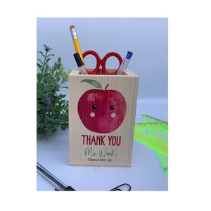 Puede incluir: Un porta lápices de madera con un diseño de manzana roja. La manzana tiene una cara sonriente y una hoja verde. El porta lápices dice "Gracias Sra. Wood Clase de 2021-22".