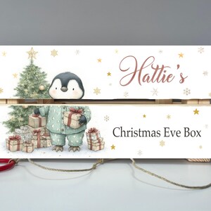 Puede incluir: Caja de Nochebuena de madera con una ilustración de acuarela de un pingüino en pijama sosteniendo un regalo. La caja está decorada con copos de nieve y un árbol de Navidad. El nombre "Hattie's" está escrito encima de "Christmas Eve Box".