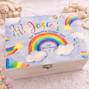 May include: A wooden keepsake box with a light blue lid featuring a rainbow, clouds, and stars design. The lid reads "Josie's Our Beautiful Flower Girl" with additional text below. The box has a silver clasp.