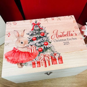 Puede incluir: Una caja rectangular de madera con una ilustración impresa de un conejo con tutú rojo junto a un árbol de Navidad decorado con adornos rojos y regalos. El texto dice Arabella's Christmas Eve box 2025 en letras rojas y negras.