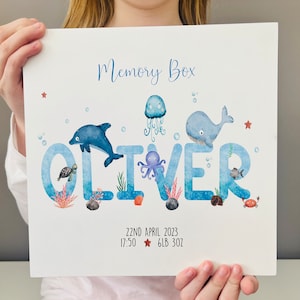 May include: A blue and white memory box print with the name "Oliver" spelled out in large letters. Each letter is decorated with a different sea creature, including a dolphin, octopus, whale, and jellyfish. The print also includes the date "22nd April 2023", the time "17:50", and the weight "6lb 3oz".